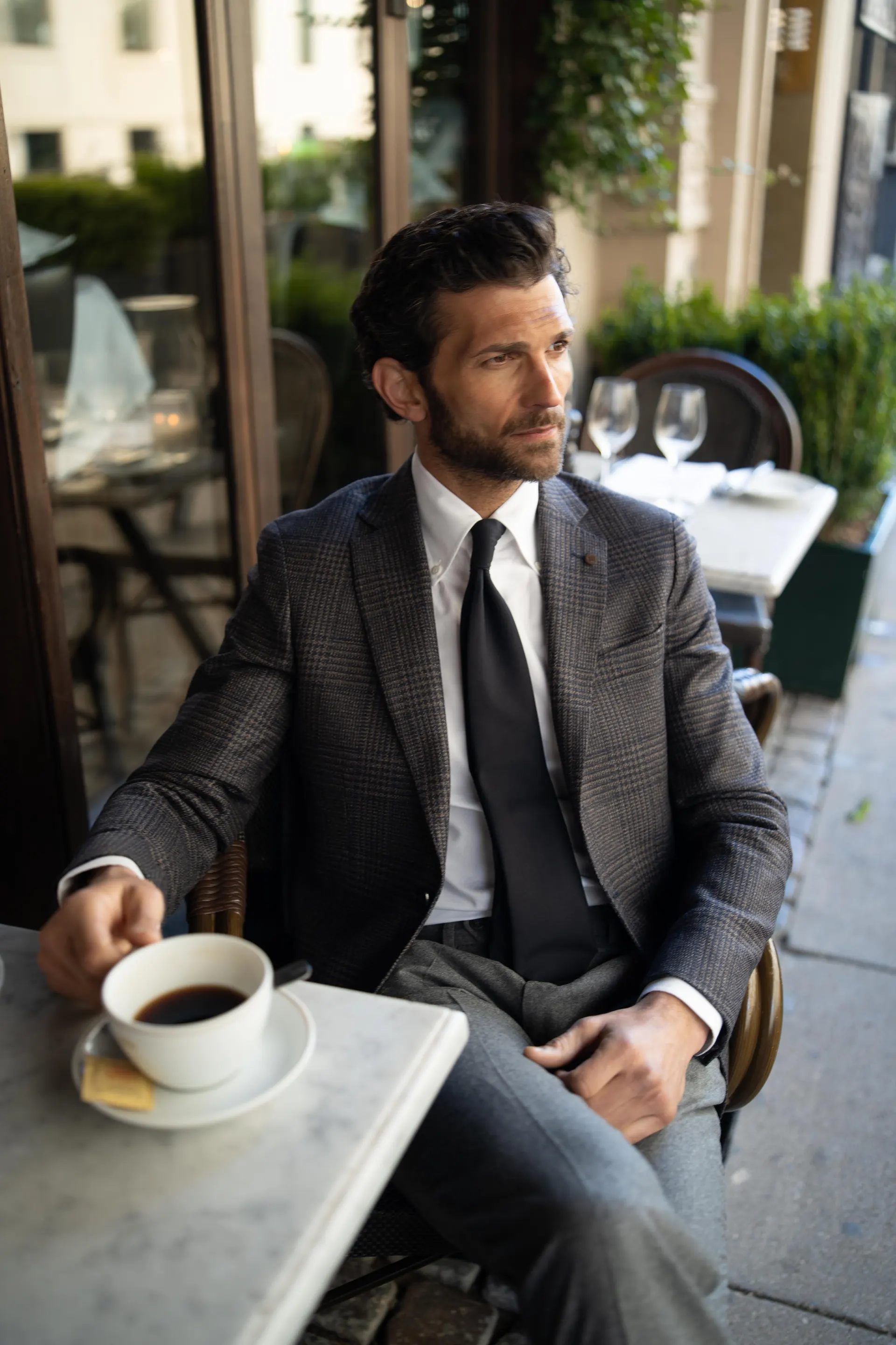 Mann i grå blazer, hvit skjorte og mørkt slips sitter ved et kafébord med en kaffekopp.
