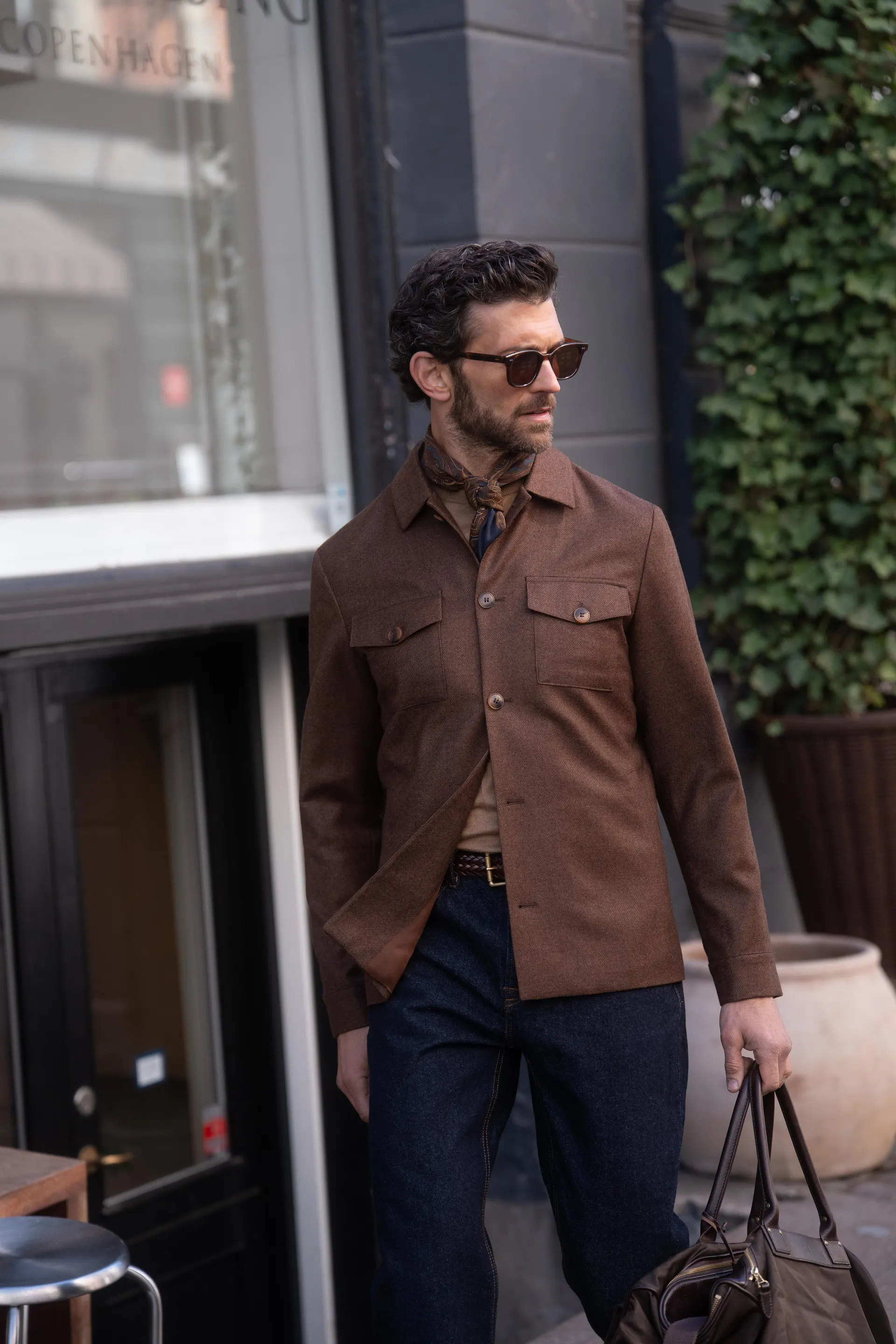 Mann i brun overshirt, mørk blå bukse og solbriller holder en weekendbag.