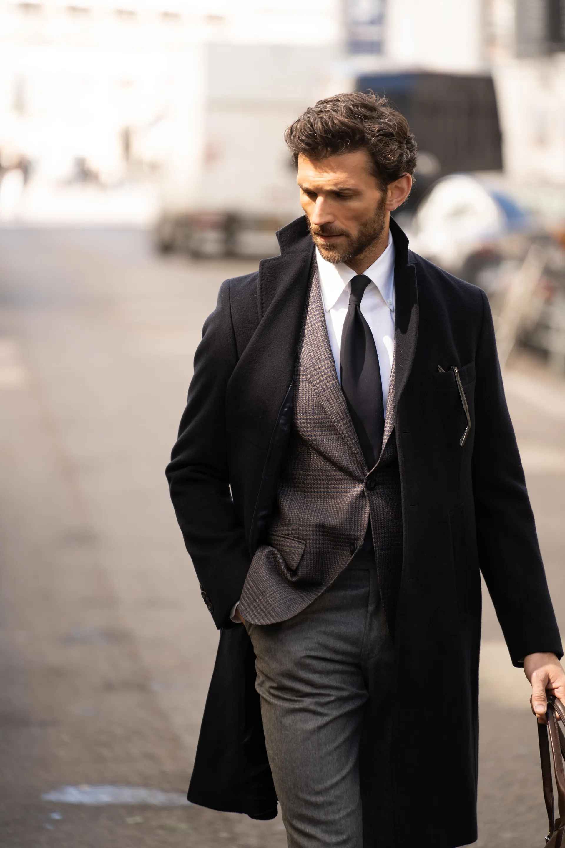 Mann i mørk frakk over gråbrun blazer, hvit skjorte og mørkt slips går langs gaten med en veske.