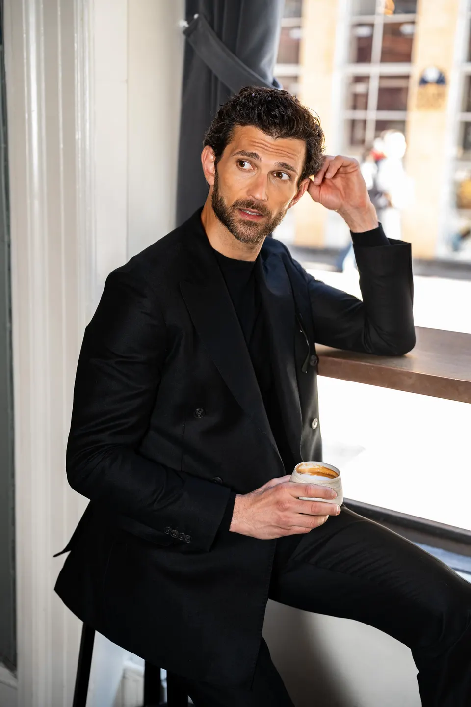 Mann i mørk dress og sort genser sitter ved et vindu med en kaffekopp.