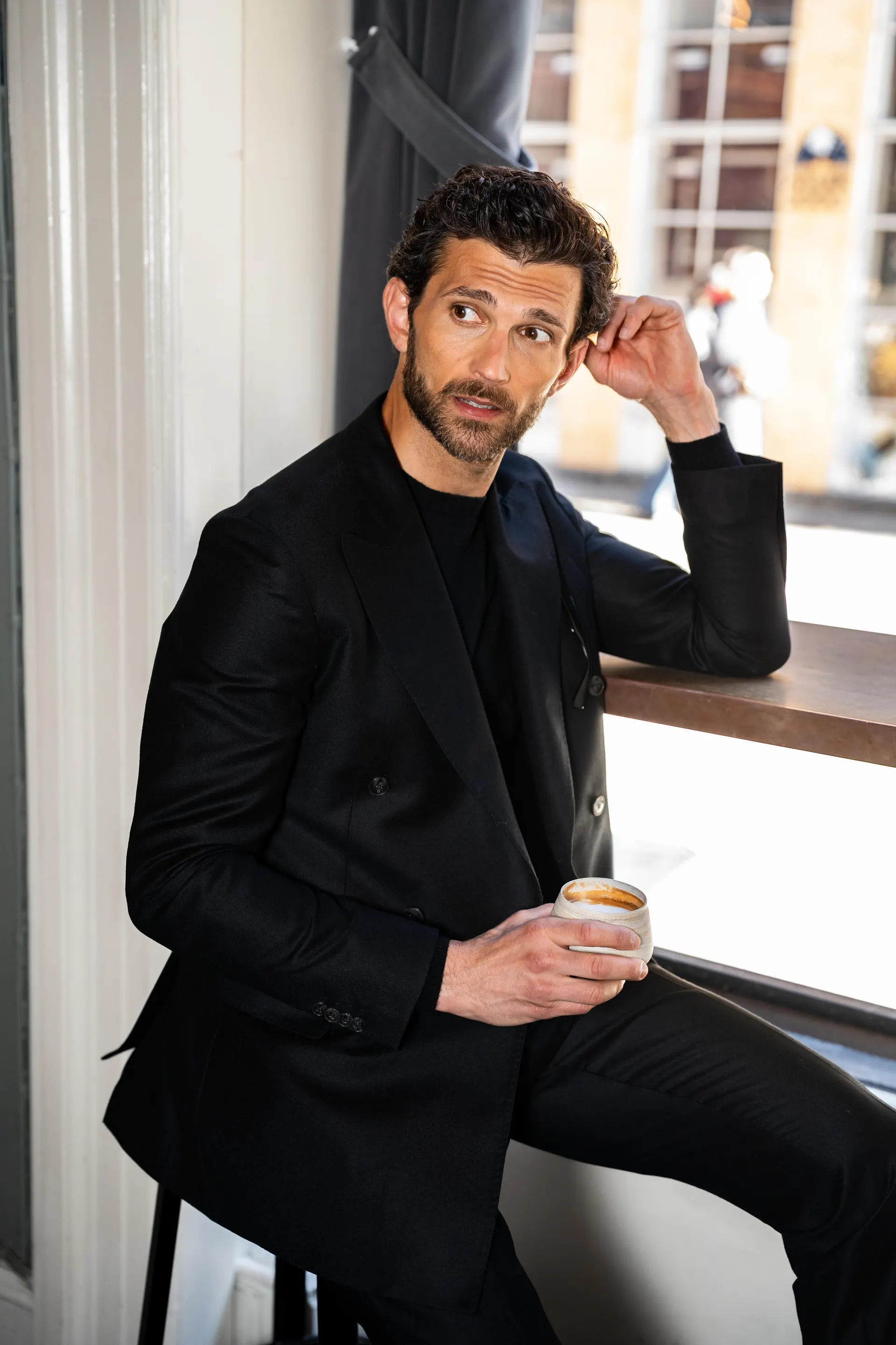 Mann i mørk dress og sort genser sitter ved et vindu med en kaffekopp.