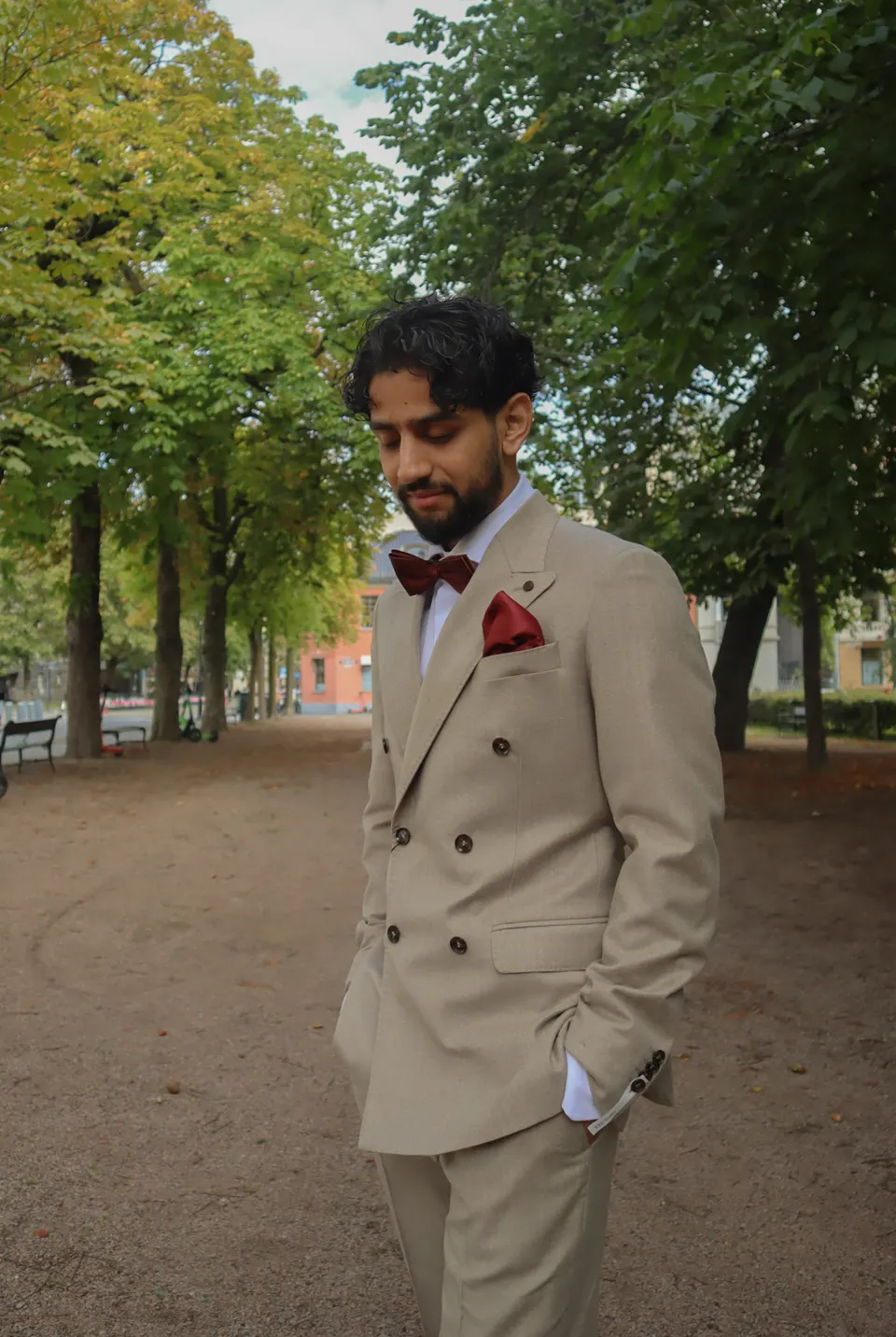 Mann i beige dobbeltspent dress med lys skjorte, burgunder sløyfe og lommetørkle, stående utendørs.
